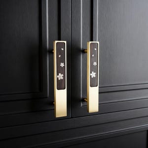 May include: Two cabinet pulls with a rectangular design. The pulls have a brass finish with a dark wood inlay. The inlay features white flower and leaf accents. The pulls are mounted on a black cabinet.