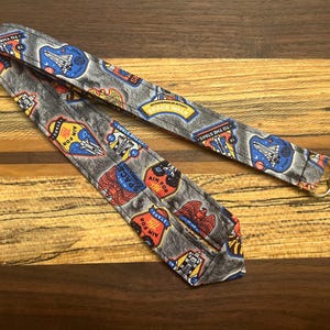 May include: A gray lanyard with a space-themed pattern featuring rockets, planets, and the text "AIM FOR THE STARS." The lanyard has a silver-colored metal clasp. The design includes red, blue, yellow, and white accents.