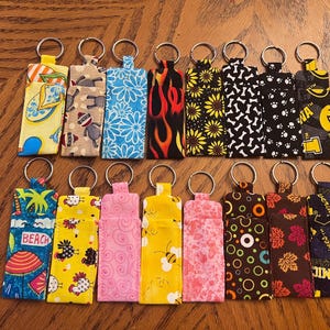 May include: A collection of fabric keychains with various patterns, including floral, animal prints, and flames. Each keychain has a metal ring and a rectangular fabric pouch. The keychains are arranged on a wooden surface.