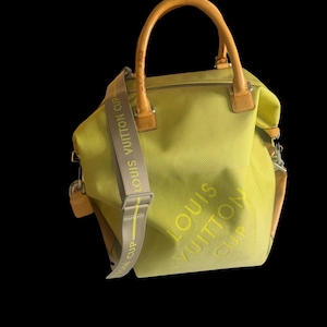 Puede incluir: Un bolso verde lima con asas y detalles en cuero marrón. El bolso tiene una correa para el hombro desmontable con el texto "LOUIS VUITTON CUP" en amarillo. El cuerpo del bolso presenta las palabras "LOUIS VUITTON CUP".