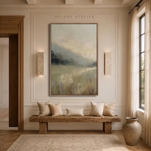 Peut inclure: Une peinture de paysage verticale encadrée représentant un champ d'herbes hautes aux tons beiges et verts doux, avec des collines sombres et brumeuses en arrière-plan sous un ciel pâle. L'œuvre est centrée au-dessus d'un banc en bois avec quatre coussins.