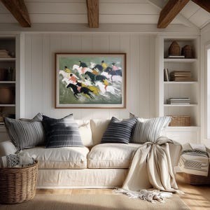 May include: A cozy living room scene featuring a cream-colored sofa with striped and solid pillows. A framed painting of horses racing hangs above the sofa. Wicker baskets and a neutral-toned rug complete the inviting space.