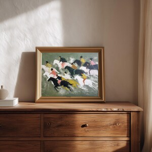 May include: A framed painting depicting a horse race with riders in various colors. The artwork is in a gold frame and rests on a wooden dresser. A small, beige decorative object sits on a white box to the left of the painting.