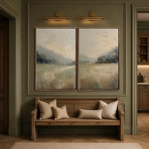 May include: A wooden bench with three beige throw pillows sits beneath two framed landscape paintings. The artwork depicts a hazy, muted field with soft hills under a pale sky, mounted on a dark olive green wall with two brass wall lights.