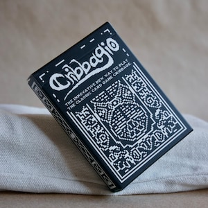 以下が含まれることがあります： 白のピクセル化されたデザインと「Cribbagio」の文字が入った黒いカードゲームボックス。テキストは「THE INNOVATIVE NEW WAY TO PLAY THE CLASSIC CARD GAME CRIBBAGE」と書かれています。ボックスは明るい色の布の上に置かれ、シンプルでクリーンな美学を作り出しています。