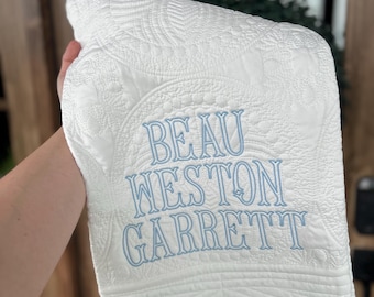 May include: A white quilted blanket featuring intricate stitched patterns and the name BEAU WESTON GARRETT embroidered in light blue thread. The fabric has a textured surface with decorative stitching throughout the entire piece.