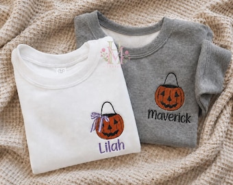 May include: A white crewneck sweatshirt with an embroidered orange jack-o'-lantern bucket featuring a purple bow and the name Lilah, and a gray crewneck sweatshirt with an embroidered orange jack-o'-lantern bucket and the name Maverick.