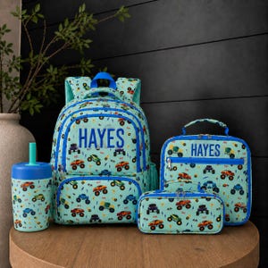 May include: A light blue backpack, lunch bag, pencil case, and tumbler set featuring a repeating pattern of colorful monster trucks and the name HAYES printed in blue on the backpack and lunch bag. Each item has blue zippers and trim.