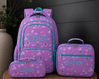 May include: A purple backpack, lunch bag, and pencil case set featuring a repeating pattern of fairies, unicorns, and castles. Each item is personalized with the name Lainey embroidered in teal thread, featuring teal zippers and trim.