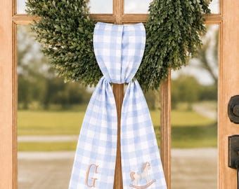 May include: A light blue and white gingham fabric ribbon tied to a green boxwood wreath. The ribbon features two long tails, one embroidered with the letter G and the other with a small rocking horse design in light brown and blue thread.