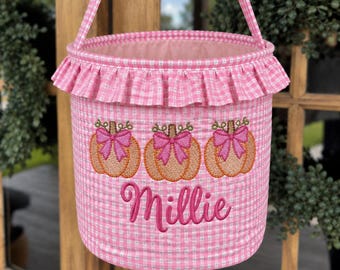 May include: A pink and white gingham fabric basket with a ruffled top edge and a matching handle. The front features an embroidered design of three orange pumpkins with pink bows and the name Millie stitched in pink cursive script lettering.
