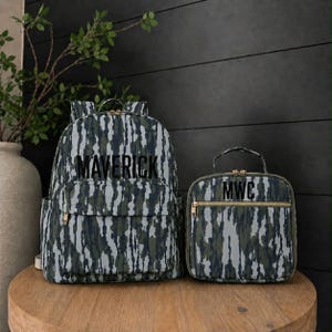 May include: A backpack and a matching lunch bag featuring a vertical camouflage pattern in shades of green, gray, and black. The backpack displays the name MAVERICK in black lettering, while the lunch bag features the initials MWC in black.