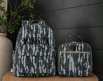 May include: A backpack and a matching lunch bag featuring a vertical camouflage pattern in shades of green, gray, and black. The backpack displays the name MAVERICK in black lettering, while the lunch bag features the initials MWC in black.