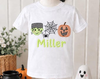May include: A white short-sleeve t-shirt featuring an embroidered Halloween design with a green Frankenstein monster head, a black spider web with a spider, and an orange jack-o'-lantern, with the name Miller stitched in lime green thread.