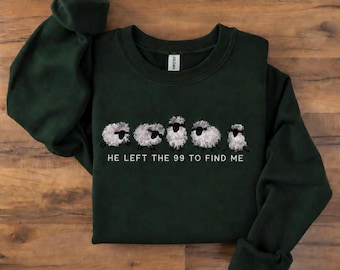 May include: A dark green long-sleeve crewneck sweatshirt featuring a graphic of five fluffy white sheep with black faces and legs. Below the sheep, the text reads HE LEFT THE 99 TO FIND ME in white capital letters. The garment has a ribbed collar.