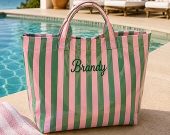 May include: A pink and green striped tote bag with the name "Brandy" embroidered in green script. The bag has matching green handles and is sitting next to a striped towel and sunglasses. The background includes a pool and ocean.