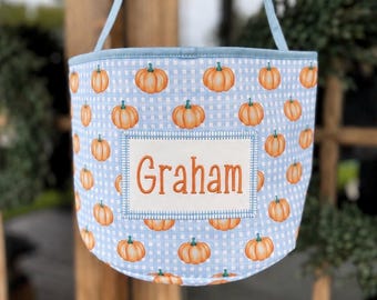 May include: A light blue and white gingham fabric bucket with a handle, featuring an all-over pattern of small orange pumpkins. A rectangular white patch is stitched to the front, displaying the name Graham embroidered in orange thread.