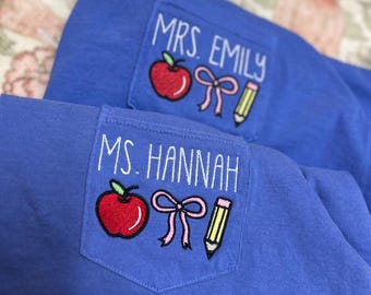May include: Two blue shirts featuring chest pockets embroidered with white text and colorful designs. One pocket reads MRS. EMILY with a red apple, pink bow, and yellow pencil. The other reads MS. HANNAH with identical apple, bow, and pencil icons.