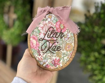 May include: A round, floral-patterned ornament with the names "Lilah Rae" embroidered in black thread. The ornament is trimmed with a gold-colored border and topped with a dusty rose-colored bow. The background is blurred, suggesting a natural setting.