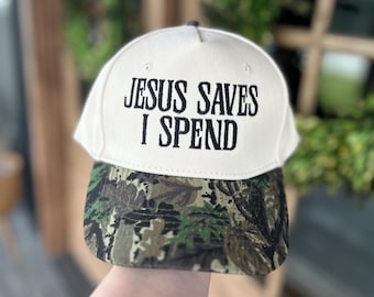 May include: A baseball cap with a cream-colored crown and a camouflage brim. The cap features the text "JESUS SAVES I SPEND" in black block letters.