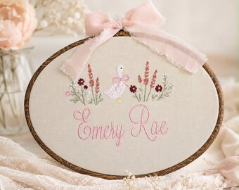 May include: Oval embroidery hoop with a cream-colored fabric featuring a pink embroidered goose, flowers, and the name "Emery Rae" in pink script. A pink ribbon bow adorns the top. The wooden hoop has a dark brown finish.