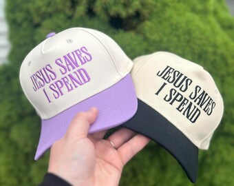 May include: Two baseball caps with the text "JESUS SAVES I SPEND". One cap has a cream-colored crown and a purple bill, the other has a cream-colored crown and a black bill. The caps are held up against a green background.