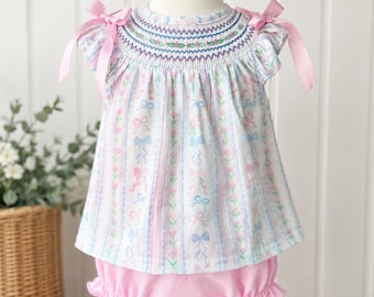 May include: A white sleeveless top with a smocked neckline featuring pink and blue floral and ribbon patterns, accented by pink ribbon bows on the shoulders, paired with solid pink ruffled bloomers on a mannequin display.
