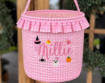 May include: A pink and white gingham fabric bucket with a ruffled top edge and a matching handle. The front features the embroidered name Millie in pink script, surrounded by small Halloween-themed icons including a witch hat, candy corn, a spider, a ghost, and a pumpkin.