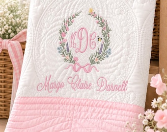 May include: A white quilted baby blanket with a pink border, featuring an embroidered floral wreath surrounding the monogram "MDC" and a small bee. The name "Margo Claire Darnell" is embroidered in pink script below the floral wreath design.