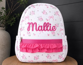 May include: A white backpack featuring a pink floral and ribbon pattern with the name Mallie embroidered in pink script. The bag includes a horizontal hot pink ribbon trim and a double-layered hot pink ruffled fabric accent above the front pocket.