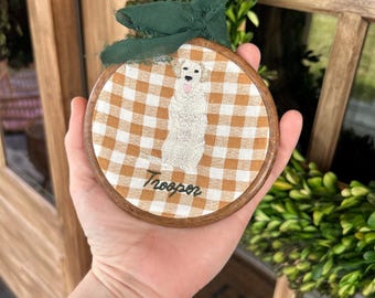 May include: Round wooden ornament with a gingham pattern and an embroidered dog design. The dog is white with a pink tongue. The name "Trooper" is written below the dog in cursive. A green ribbon is attached at the top.