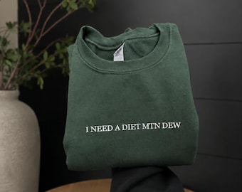 May include: A dark forest green crewneck sweatshirt featuring the white embroidered text "I NEED A DIET MTN DEW" centered on the chest. The garment has a ribbed collar and long sleeves, showcasing a simple design with clear, legible lettering.