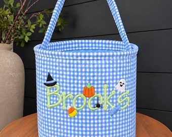 May include: A cylindrical fabric bucket with a blue and white gingham pattern and a matching handle. The front features the name Brooks embroidered in light green, decorated with small Halloween icons including a witch hat, pumpkin, ghost, and candy.