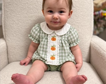 May include: A light green and white gingham romper featuring a white Peter Pan collar and a central vertical panel decorated with three embroidered orange pumpkins. The garment has short puff sleeves and a small orange bow hair accessory.