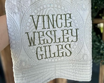 May include: A white quilted fabric square featuring intricate circular stitching patterns and the embroidered dark green text VINCE WESLEY GILES centered on the front. The fabric has a soft texture with detailed geometric quilting designs.