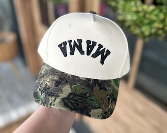 May include: Cream-colored baseball cap with the word "MAMA" embroidered in black. The brim features a camouflage pattern in shades of green, brown, and tan. A casual accessory for everyday wear.