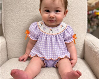 May include: A light purple and white gingham baby outfit featuring a smocked collar with embroidered ghost and bow motifs. The top has short puff sleeves with orange ribbon ties, a scalloped hem with orange trim, and matching purple bloomers.