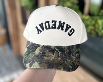 May include: A white baseball cap with the word "GAMEDAY" embroidered in black across the front. The brim of the cap features a camouflage pattern in shades of green, brown, and tan.