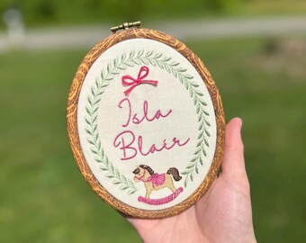 May include: An oval embroidery hoop with a faux wood grain frame containing off-white fabric. The fabric features a green embroidered laurel wreath, a pink bow, the name Isla Blair in pink cursive script, and a small rocking horse graphic.