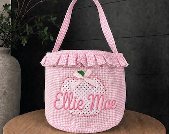 May include: A pink and white gingham fabric basket featuring a ruffled top edge and a single handle. The front displays an embroidered white apple shape with a small pink bow and the name Ellie Mae stitched in pink cursive lettering.