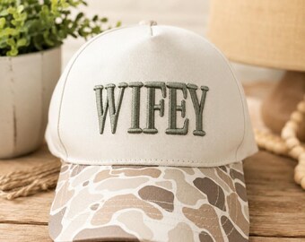 May include: A cream-colored baseball cap with the word "WIFEY" embroidered in green thread. The brim features a camouflage pattern in shades of brown, tan, and gray. The hat is a casual accessory.