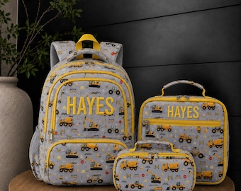 May include: A gray backpack, lunch bag, and pencil case set featuring a yellow construction vehicle pattern. Each item displays the name HAYES in yellow block letters. The set includes yellow zippers and trim, with a top handle on the bag items.