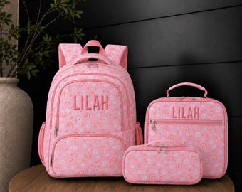 May include: A pink backpack, lunch bag, and pencil case set featuring a repeating pattern of small rainbows and white flowers. Each item is personalized with the name LILAH embroidered in a matching pink thread on the front pocket.
