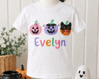 May include: A white short-sleeved t-shirt featuring an embroidered design of a pink pumpkin, a purple pumpkin with bolts, and an orange cat face. Below the pumpkins, the name Evelyn is embroidered in multicolored, textured, block-style letters.