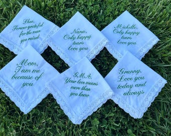 May include: Six white handkerchiefs with green embroidered text and lace trim are arranged on green grass. The handkerchiefs have various messages, including "Forever grateful," "Only happy tears love you," and "Love you today and always."