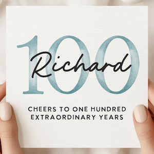 May include: A square card with the number "100" in teal watercolor and the name "Richard" in black script. The text "CHEERS TO ONE HUNDRED EXTRAORDINARY YEARS" is below. The card is held by hands with manicured nails.