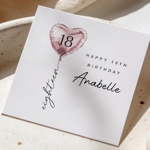 May include: A white square birthday card with a heart-shaped balloon illustration. The balloon is pink and features the number 18. The card includes the text "Happy 18th Birthday Anabelle" in black script.