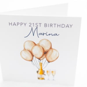 May include: A white greeting card with the text "HAPPY 21ST BIRTHDAY Marina" in navy blue. The card features a watercolor illustration of champagne bottles, glasses, and balloons in shades of gold and beige.