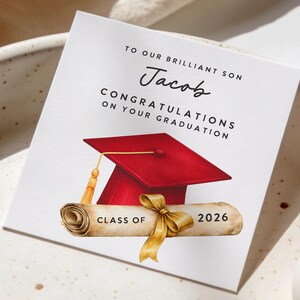 May include: A graduation card with the text "TO OUR BRILLIANT SON Jacob CONGRATULATIONS ON YOUR GRADUATION". The card features a red graduation cap, a scroll with a gold ribbon, and the text "CLASS OF 2026".