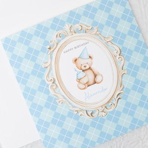 May include: A birthday card with a light blue diamond pattern background. The card features a decorative frame with a teddy bear wearing a party hat and holding a cupcake. The words "Happy Birthday" and a name are written on the card.